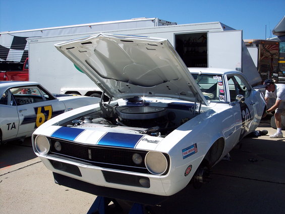 White 1969 Chevrolet Camaro race car with blue stripes, hood open, on jack stands. Another Camaro with number 67 visible