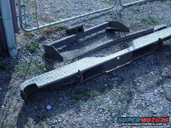 Two metal bumpers and a hitch receiver sit on gravel near a fence