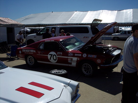 Red race car with number 70 and 'STARK HICKEY FORD Inc.' text, hood open. White car in foreground. Tents and people in background