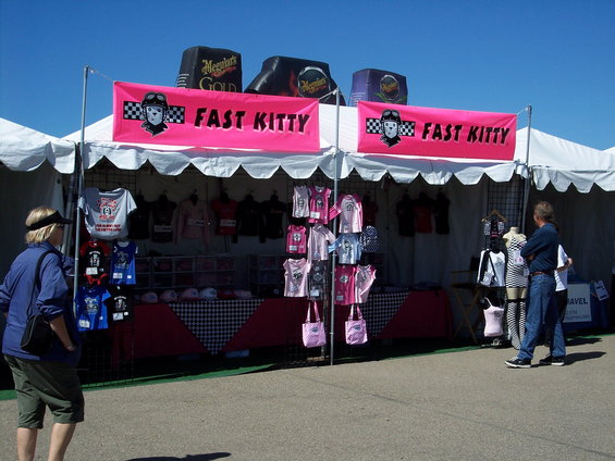 Tents with pink banners reading "FAST KITTY" display t-shirts and bags under a clear blue sky