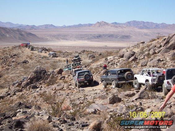 Line of off-road vehicles, including a dark Bronco, driving on a rocky desert trail