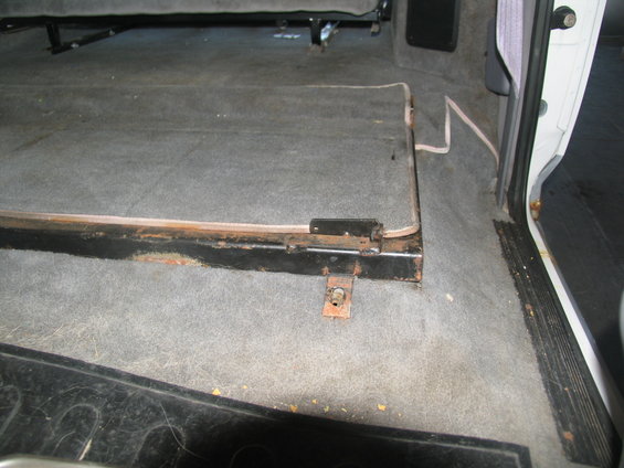 Interior shot of a vehicle's carpeted floor with a folded-down seat base and metal frame. Rust is visible on the frame