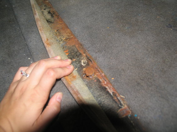 Rusted metal trim with exposed fasteners on gray carpeted floor. A hand with a ring is visible