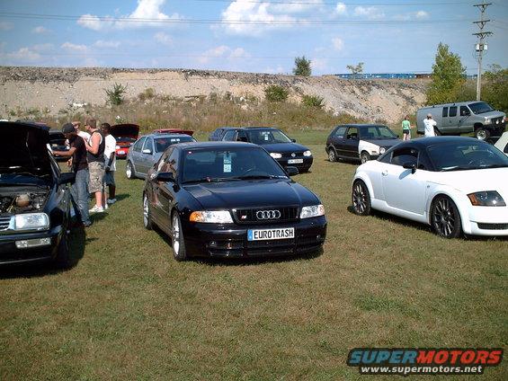 Black Audi S4 with 'EUROTRASH' plate, parked on grass with other cars and people