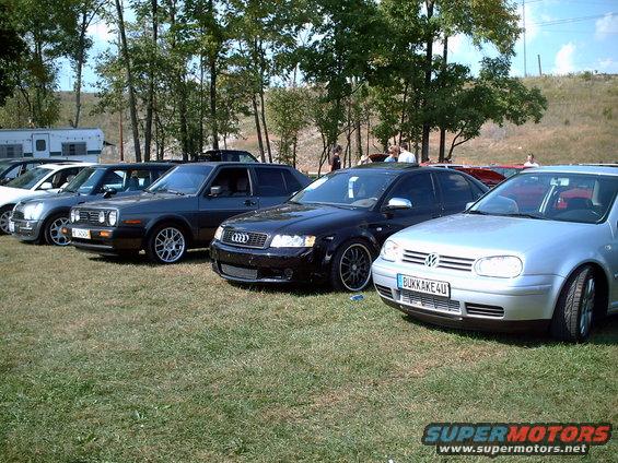 Several cars parked in a row on a grassy field with trees in the background. A black Audi and a silver Volkswagen are in the foreground