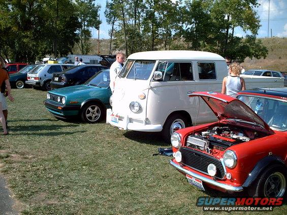 Cream-colored Volkswagen Type 2 bus parked on grass, front quarter view, hood open on a red car in foreground