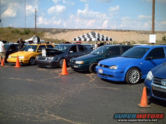 Several Volkswagen cars, including a blue Golf and a dark gray sedan, parked in a lot under a checkered tent