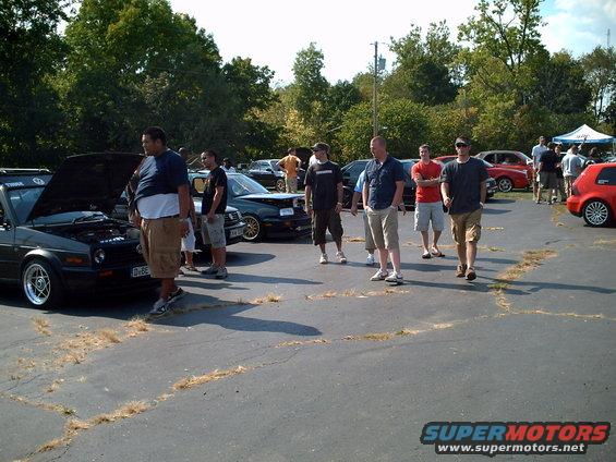 People milling around a car meet, with several cars visible including a black hatchback with its hood up and a red hatchback