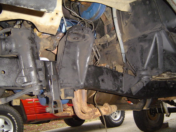 Underside of a vehicle showing suspension and exhaust components. A red vehicle is partially visible in the background