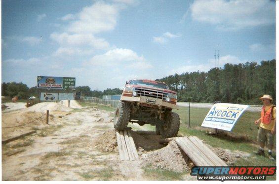 red lifted pickup truck driving over wooden ramps on dirt course, person in orange vest nearby