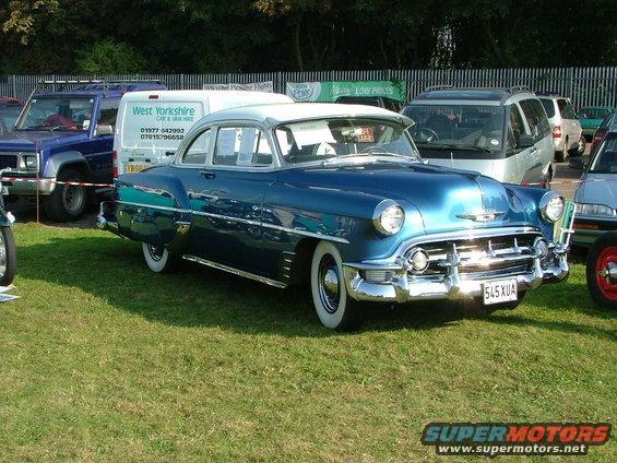 Blue classic sedan with white roof and whitewall tires. License plate reads 545 XUA. Other vehicles and a van are in the background