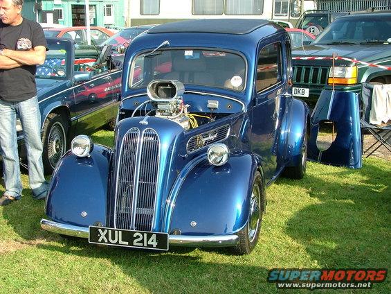 Blue hot rod with exposed engine and custom grille. License plate reads XUL 214. Other vehicles and a person are visible in the background