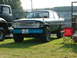 1988 Ford Sport - This modified sedan features large off-road tires and a bright blue bumper, parked on grass next to another vehicle.