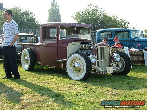 Maroon hot rod pickup truck with exposed engine and whitewall tires parked on grass. Another blue vehicle is behind it