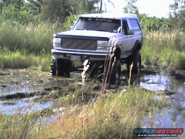 White SUV with lifted suspension driving through a muddy field with tall grass