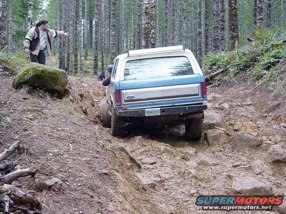 Blue and white Ford Bronco with a silver stripe on the rear, navigating a muddy off-road trail