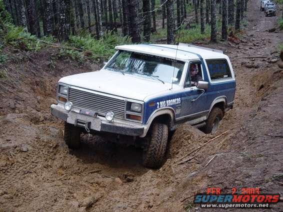 Blue and white Ford Bronco stuck in mud on a wooded trail. Text on door reads '2 BIG BRONCOS'. Another vehicle is visible further up the trail