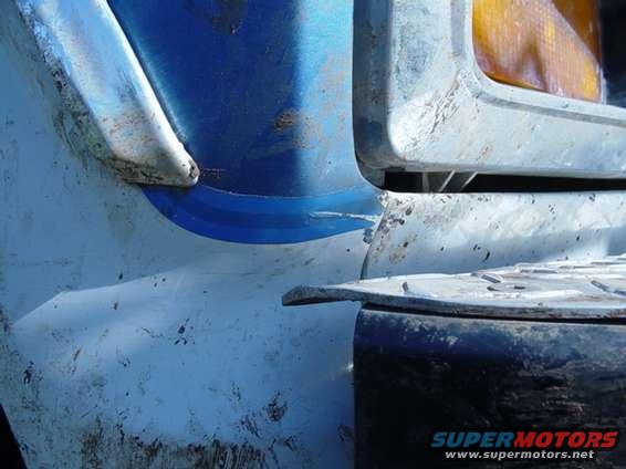 Damaged front corner of a blue and white vehicle, showing a dented fender and headlight housing