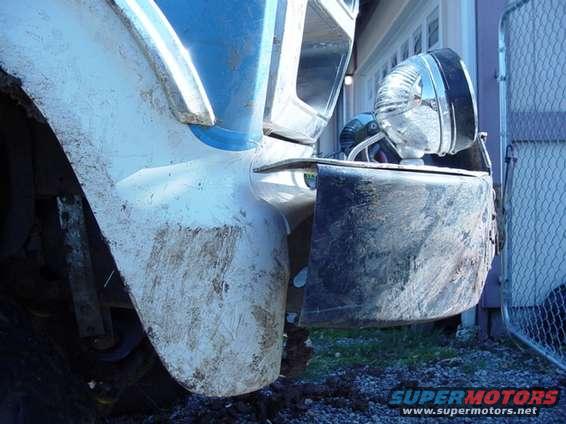 Front fender and bumper of a truck, covered in mud. A chrome headlight is visible