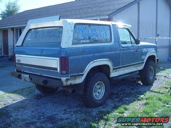 Blue and white Ford Bronco parked outside a house. It has a dented rear bumper and dirty windows