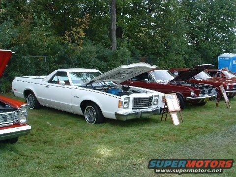 White Ford Ranchero with hood open, parked on grass next to other cars. Other vehicles are visible in the background