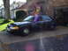 2003 Ford Crown Victoria - A black sedan is parked in a driveway in front of a house and an open garage.