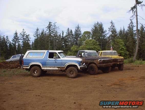 Blue and white Ford Bronco and two Ford F-Series trucks parked on dirt with trees in background