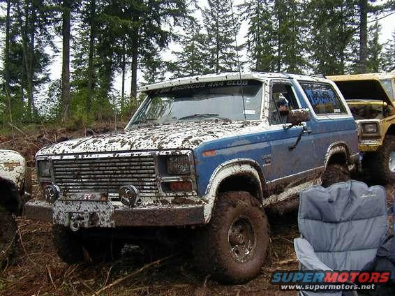 Mud-covered blue and white Ford Bronco with large tires, parked in a wooded area next to a yellow vehicle