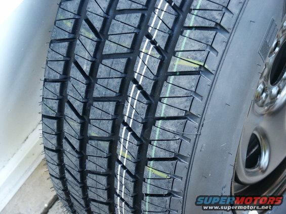 Close-up of a tire with a distinct tread pattern and white and green markings. A silver rim is visible on the right