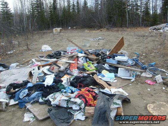 Pile of garbage and debris including clothes, boxes, and a blue bicycle in a wooded area