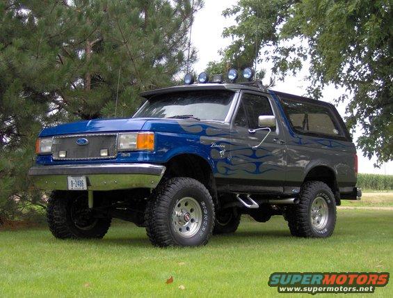 Blue and gray Ford Bronco with flame decals, lifted with large tires. It has a visor with lights on top