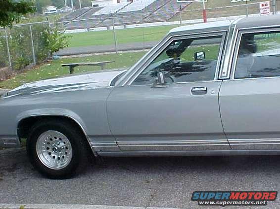 Side view of a silver 1980s Ford sedan with aftermarket wheels. Stadium seating is visible in the background