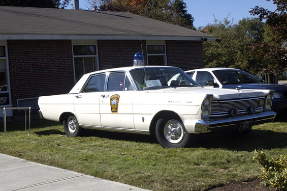 White 1965 Ford sedan police car, front quarter view, parked on grass. It has a blue light on the roof and a police badge on the door