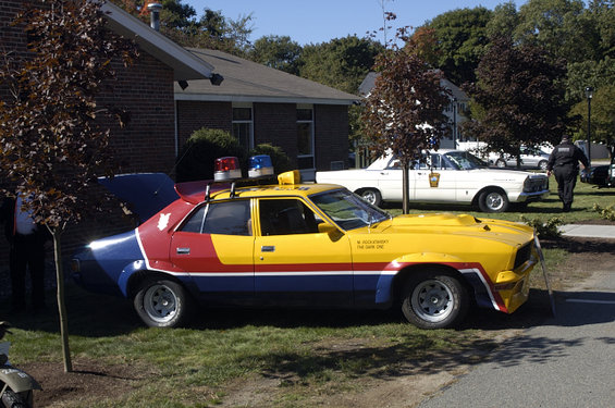 Yellow and blue police car with red stripe, light bar, and custom hood scoop. White sedan in background