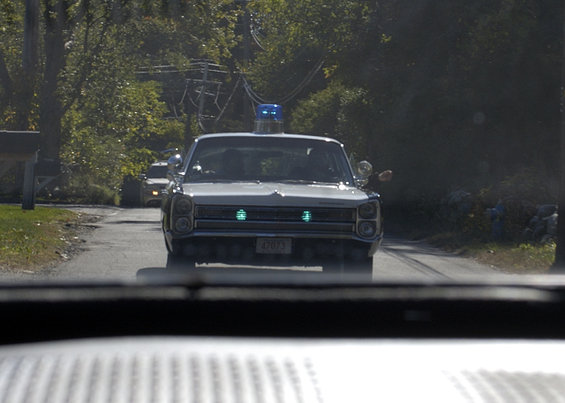 White police car with blue light bar and green headlights, driving on a road. License plate reads 42873