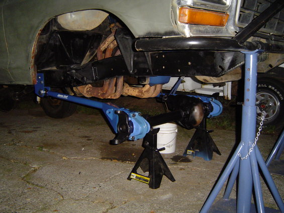 Vehicle front axle and suspension components are blue and black, with rusty exhaust manifolds visible. The vehicle is supported by jack stands