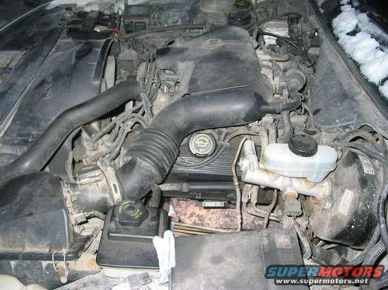 Engine bay with black air intake hoses, oil cap, and brake fluid reservoir. Some snow is visible on the windshield