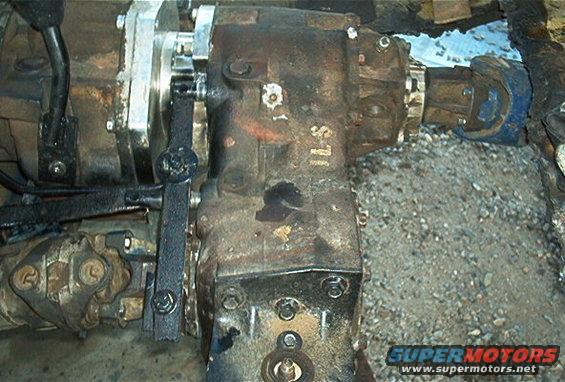 Close-up of a rusty transfer case with a black shifter lever and linkage. Gravel is visible in the background