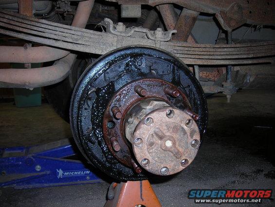 Rear drum brake assembly on a vehicle supported by an orange jack stand. Leaf springs and exhaust pipes are visible above