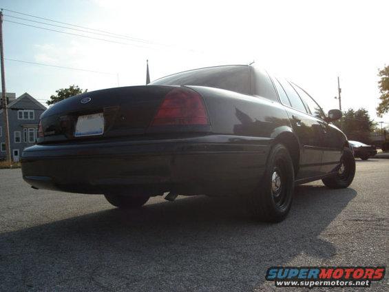 Rear quarter view of a black sedan parked on asphalt. Another car is visible in the distance