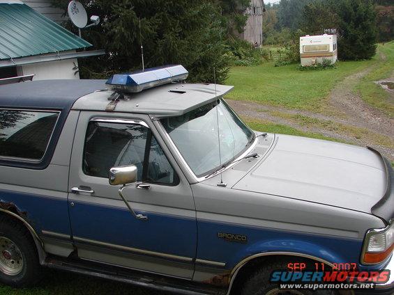 1992 Ford Bronco with a blue light bar on the roof, parked in a grassy area near a barn and a camper
