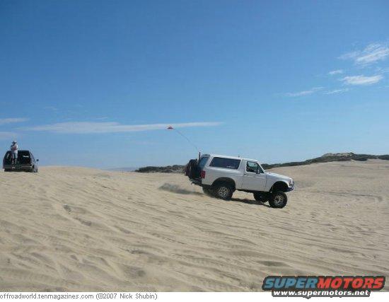 White 1994 Ford Bronco driving on sand dunes with a person in the back of another vehicle