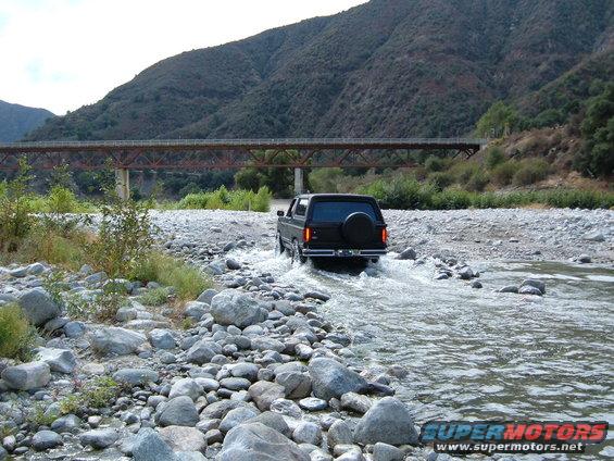 Black Ford Bronco driving through a rocky riverbed under a bridge