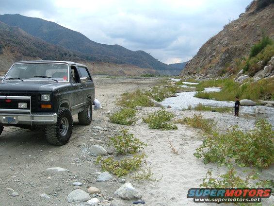 Black Ford Bronco parked on dirt near a dry riverbed with mountains in background