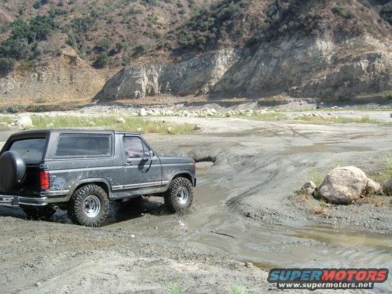 Dark gray Ford Bronco driving through mud, spare tire on rear door