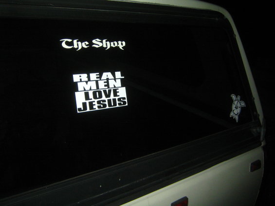 White truck's rear window with 'The Shop' and 'REAL MEN LOVE JESUS' stickers