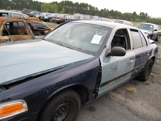 Police car with blue and light blue paint, hood open, in a junkyard with other cars