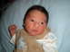 2005 Mazda 6 - A close-up of a baby's face and upper body, lying on a blue blanket. The baby is wearing a brown vest over a patterned onesie.