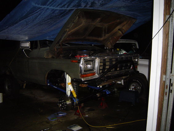 Olive green 1979 Ford truck with hood up, undergoing front suspension work, parked under a blue tarp