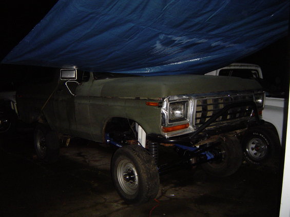 Modified pickup truck with custom front bumper and lifted suspension, partially covered by a blue tarp. Another white truck is visible behind it